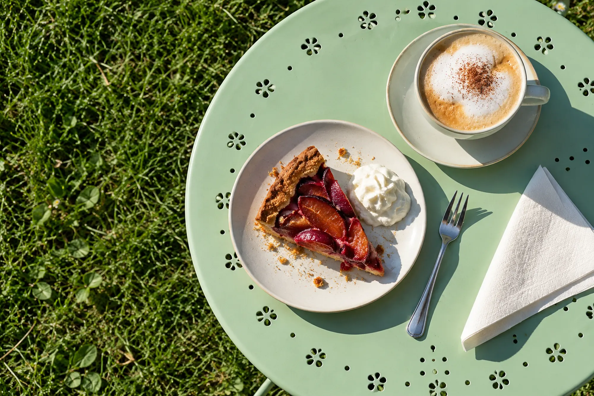 Von oben: Obstkuchen mit Sahne, Kuchengabel, Cappuccino und Serviette auf einem mintgrünen Gartentisch im Gras, harte Nachmittagsschatten (KI-Illustration)